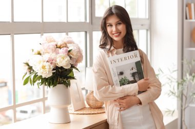 Evde elinde dergi ve şakayık çiçekleri olan genç bir kadın var.