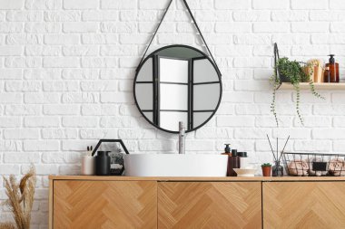 Beyaz tuğla duvarın yanında lavabo kasesi ve banyo aksesuarları olan tahta bir dolap.