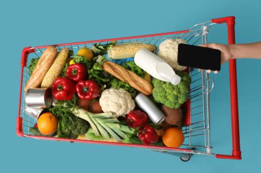 Cep telefonlu bir kadın mavi arka planda yiyecek dolu alışveriş arabalarını itiyor.