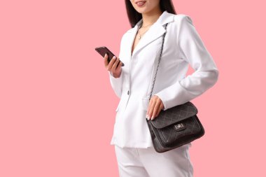 Pembe arka planda şık çantalı ve cep telefonlu güzel bir Asyalı kadın, yakın plan.