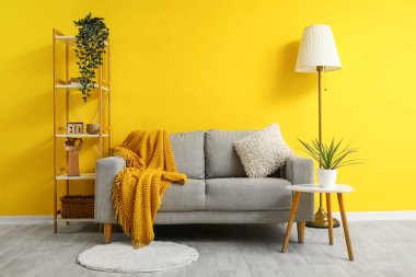 Ekose ve yastık gri kanepede, lamba, masa ve ahşap askı sarı duvarın yanında