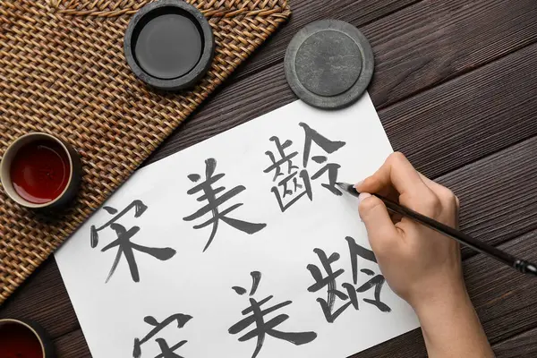 Asyalı hiyeroglifler, fırça, mürekkep ve ahşap arka planda çay fincanları olan kadın eli, yakın plan. Uluslararası Haiku Şiir Günü