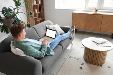 Genç adam evdeki kanepede dizüstü bilgisayar kullanıyor.