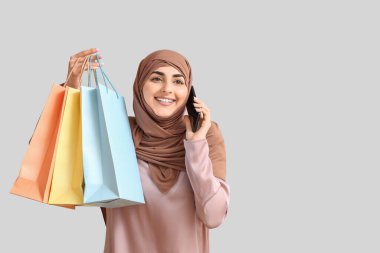 Alışveriş çantalı genç Müslüman kadın gri arka planda telefonla konuşuyor.
