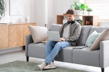 Erkek öğrenci evdeki kanepede dizüstü bilgisayarla ders çalışıyor.