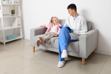 Panosu olan bir erkek çocuk doktoru ve klinikteki kanepede oturan küçük bir kız.