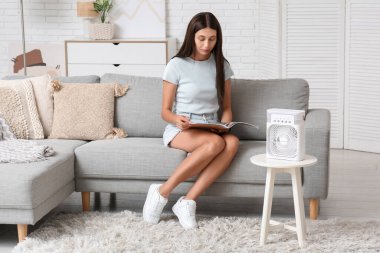 Oturma odasında nemlendiricisi olan taşınabilir yelpazenin yanında dergi okuyan güzel bir kadın.