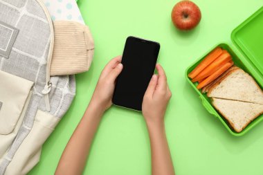 Beslenme çantasının yanında cep telefonu ve yeşil arka planda sırt çantası olan çocuk elleri. Üst görünüm