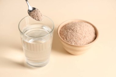 Bej arka planda psyllium kabuklu bir bardak su ve kaşık.