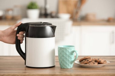 Modern elektrikli su ısıtıcısı, bir fincan sıcak çay ve mutfaktaki ahşap masada tatlı kurabiyeler.