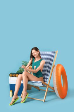 Mavi arka planda plaj aksesuarlarıyla iskemlede güneş kremi süren genç bir kadın.