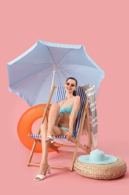 Güverte sandalyesinde pembe arka planda plaj şemsiyesi olan genç bir kadın.