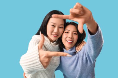 Güzel, genç, mutlu Asyalı kız kardeşler mavi arka planda fotoğraf çerçevesi yapıyorlar.