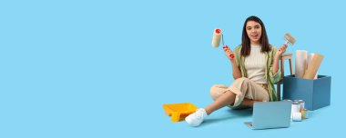 Mavi arkaplan üzerine boya silindiri, fırça, dizüstü bilgisayar ve duvar kağıtlarıyla genç bayan iç mimar.