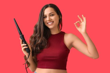Beautiful young woman with curling iron showing OK on red background