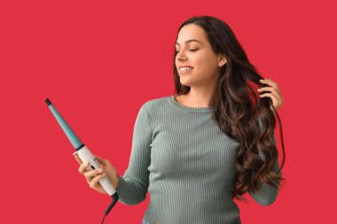 Beautiful young woman with curling iron on red background