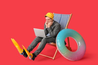 Şişirilebilir yüzüklü ve yüzgeçli bir ofis çalışanı kırmızı arka planda dizüstü bilgisayarıyla güverte koltuğunda oturuyor.