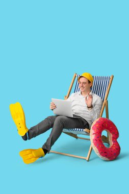 Ofis çalışanı dizüstü bilgisayarla güvertede oturuyor ve mavi arka planda tatilin hayalini kuruyor.