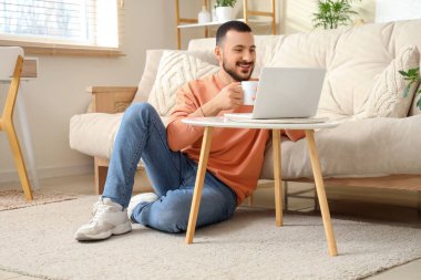 Oturma odasında dizüstü bilgisayar kullanarak çay içen yakışıklı genç adam.