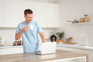 Genç adam mutfakta dizüstü bilgisayar kullanarak bir fincan sıcak çay içiyor.