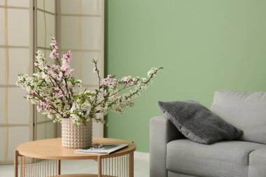 Oturma odasındaki yeşil duvara yaslanmış sehpanın yanında dergi bulunan çiçek açan bir vazo.