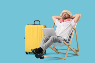 Dizüstü bilgisayarı güverte koltuğunda ve mavi arka planda bavulu olan genç bir iş adamı. Yaz tatili kavramı