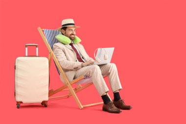 İşadamı kırmızı arka planda bavuluyla güverte koltuğunda dizüstü bilgisayar kullanıyor. Yaz tatili kavramı