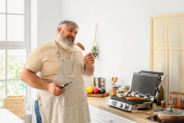 Tong pişiren olgun bir adam. Lezzetli sosisler ve sebzeler. Mutfaktaki modern elektrikli ızgaranın üstünde.