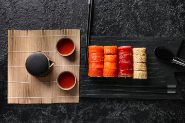 Lezzetli suşi ruloları, yemek çubukları, çaydanlık ve siyah grunge arka planda bir fincan çay.