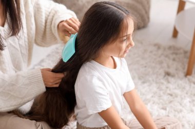 Genç anne, evde yerde otururken kızının saçını tarıyor.