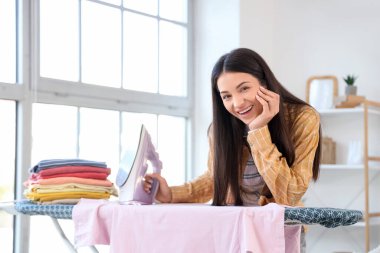 Evde demir ve giysi yığınıyla güzel, mutlu bir kadın var.