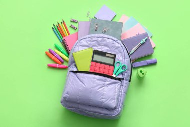 Yeşil arka planda farklı kırtasiye malzemeleriyle leylak rengi okul çantası.