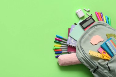 Değişik kırtasiye malzemeleri ve yeşil arka planda modern kulaklıklar olan şık bir okul çantası.