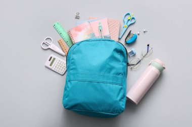 Üzerinde farklı kırtasiye malzemeleri ve gri arka planda su şişesi olan mavi okul çantası.