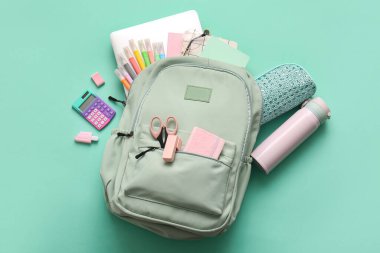 Farklı kırtasiye malzemeleri ve turkuaz arka planda su şişesi olan şık bir okul çantası.