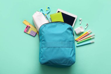 Mavi okul sırt çantası, farklı kırtasiye malzemeleri, kulaklıklar ve turkuaz arka planda modern tablet bilgisayar.
