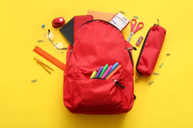Kırmızı okul sırt çantası, farklı kırtasiye malzemeleri ve sarı arka planda gözlük.