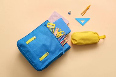 Bej arka planda farklı kırtasiye malzemeleriyle mavi okul çantası.