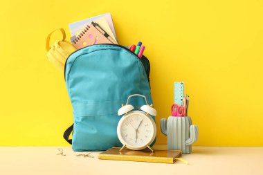 Sarı zemin üzerinde alarm saati ve kırtasiye malzemesi olan okul çantası.