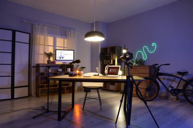 Karanlık podcaster stüdyosunun içinde iş yeri, raf üniteleri ve parlayan lambalar var.