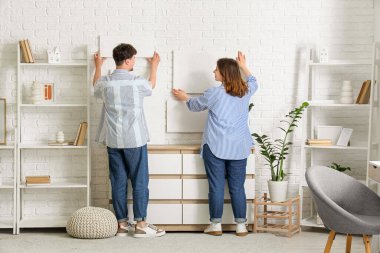 Genç bir çift evdeki beyaz tuğla duvara resim asıyor.