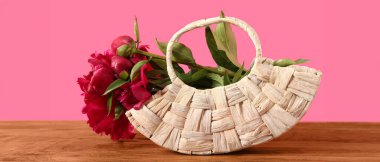 Ahşap masada pembe arka planda güzel şakayık çiçekleri olan hasır çanta.