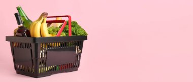 Farklı taze ürünlerle dolu siyah alışveriş sepeti ve pembe arka planda muzlar.