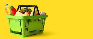 Farklı taze ürünlerle dolu yeşil alışveriş sepeti ve sarı arka planda bir şişe yağ.