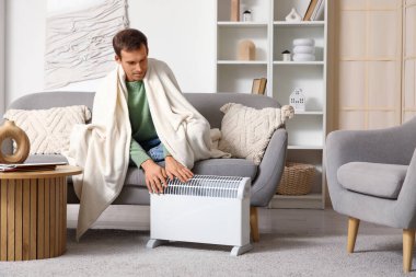 Ekoseli genç adam evdeki elektrikli konteyner ısıtıcısının yanında ellerini ısıtıyor.