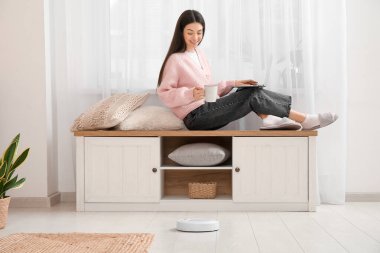 Çay fincanı kullanan genç bir kadın bankta dizüstü bilgisayar ve elektrikli süpürge kullanıyor.