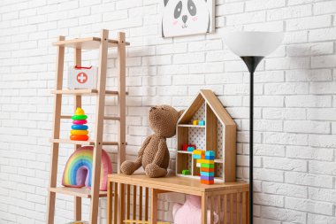 Çocuk odasının içinde raflarda oyuncaklar ve beyaz tuğla duvarın yanında lamba var.