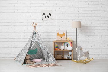 Raflarda bir sürü oyuncağı olan çocuk odasının içi. Çadır, sallanan at ve lamba, beyaz tuğla duvarın yanında.