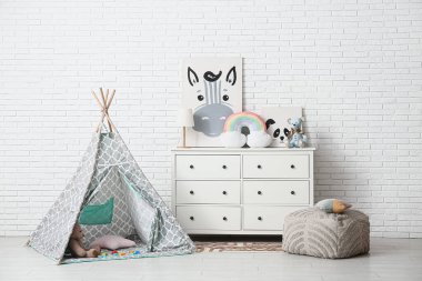 Beyaz tuğla duvarın yanında oyun çadırı, bir sürü oyuncak ve lamba olan çocuk odasının içinde.