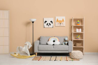 Çocuk odasının içinde kanepe, lamba, raflarda bir sürü oyuncak ve bej duvarın yanında sallanan at var.